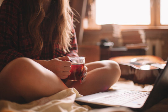 Young female holding cup of tea while sitting on bed with laptop