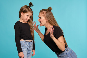 Mom and daughter with a funny hairstyles, dressed in black shirts and blue denim jeans are posing against a blue studio background. Close-up shot.