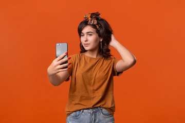 Fototapeta premium Pretty brunette woman in a t-shirt and beautiful headband making selfie with spartphone standing isolated over orange background. Mobile photography