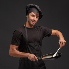 Young male cook in a black apron and hat cooks vegetables in a pan.