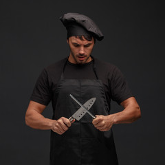 Young male cook in a black apron and hat sharpening a knife.