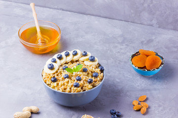 Oatmeal, honey and fruit for breakfast. Oatmeal with blueberries, banana and mint. Healthy porridge with cereal flakes, dried apricots and nuts. Cooked oatmeal in bowl on cement