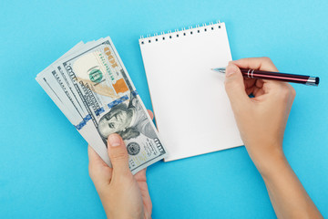 A woman holds money in her hand and writing in a notebook on a blue background. Concept of planning of the budget. Blank white notebook with copy space for your ideas.