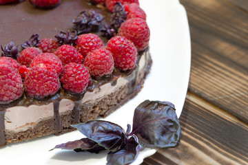 Tasty and harmless dessert background. Vegan chocolate cake with raspberries on white plate with basil leaves. Wooden table. Top angle view.