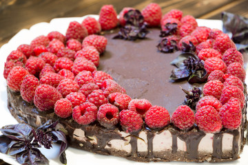 Sweet healthy present for lovers. Close up raw foods chocolate cake with raspberries on white plate with basil leaves. Cooked in the shape of heart