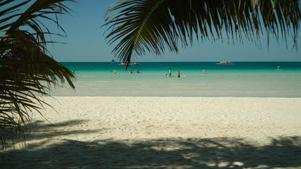 Tropical beach with tourists and clear blue sea through palm branches. Summer and travel vacation...