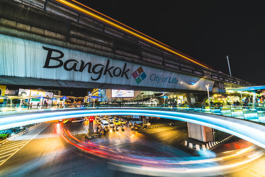 Bangkok, Thailand - Nov 25, 2017: Traffic light trail at road junction near MBK shopping center in Bangkok, Thailand. Tourism advertising logo on BTS sky train track, pedestrain walks on skywalk area