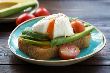 A piece of grilled white bread with poached egg , fresh basil leaves on a blue plate and small tomatoes. Front view. 