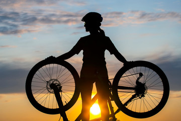 Obraz premium Silhouette of a girl cyclist with a bicycle on the background of a sunset sky. Healthy lifestyle and sport. Leisure and hobbies