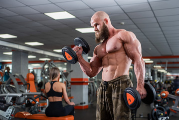 Brutal, muscular, bald with a beard athlete in the gym. Lifts heavy weights, trains the biceps....