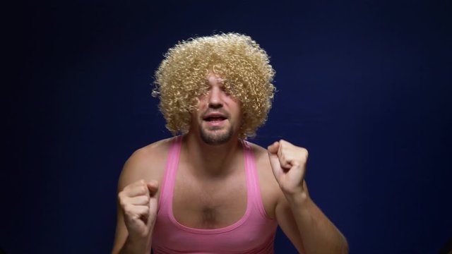 crazy handsome young guy in a curly wig and a pink t-shirt against a dark background is dancing funny, shows his muscles