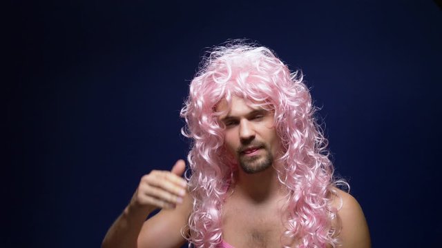 crazy handsome young guy in a curly wig and a pink t-shirt against a dark background is dancing funny, shows his muscles
