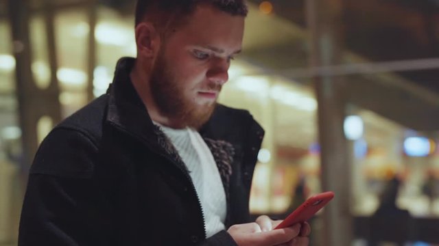 Young nice-looking man browsing application on smartphone exploring social media walking along modern glass building in city center. Night time. People and technology.