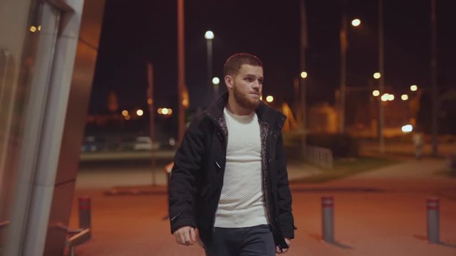 Attractive young bearded man in stylish outfit walking in the street wof beautiful modern downtown at deep night. Confident person portrait. Night cityscape.