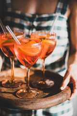 Young woman in checkered dress holding tray with glasses of Aperol Spritz aperitif cold drink with...