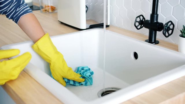 Close-up of female hand in rubber glove washing sink with wet cloth doing housework in kitchen caring for hygiene. Apartment, housekeeping and cleaning service concept.