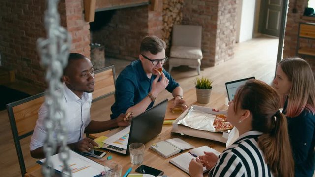 Business Team Enjoy Eating Lunch Together at Workplace with Laptops. Diverse Group of Four People Eats Pizza and Discusses Project in Loft Office. 4K High Angle Gimbal Circling Medium Shot
