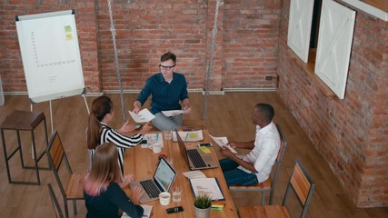 Top View of Teamwork of Diverse Group of People. Young Business Man Team Leader is Giving Papers to Employees at Meeting in Startup Loft Office. 4K Gimbal Overhead Panning Shot in Slow Motion