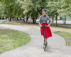 A young woman with purple hair rides an electric scooter in a park. A stylish girl with a shaved temple in a plaid shirt, a long red skirt and a bow tie is riding around the city on a modern device. 