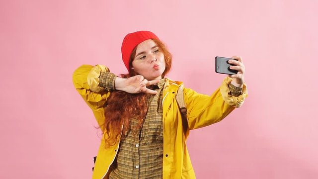 Young charming red-haired girl takes pictures of herself on the phone. Studio shot isolated on pink