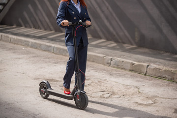 Obraz premium A red-haired girl in a jacket and trousers drives an electric scooter along the wall. A business woman in a pantsuit and red high heels rides around the city in a modern car. Office worker. Dress code