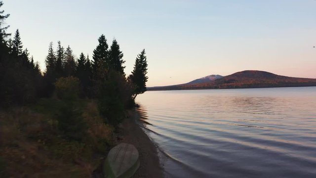 Aerial view of Zuratkul park; drone flys over mountain lake along coast with boats in early morning; snow tops, Ural mountain ridges on background; reflection of colorful sunrise sun on water surface
