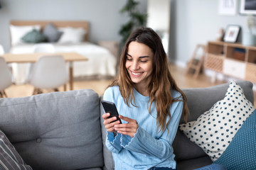 Cheerful young woman using mobile phone while sitting on a couch at home. - Powered by Adobe