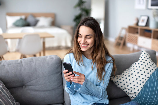 Cheerful young woman using mobile phone while sitting on a couch at home.
