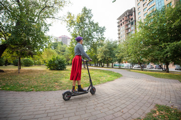 A young woman with purple hair rides an electric scooter in a park. A stylish girl with a shaved...