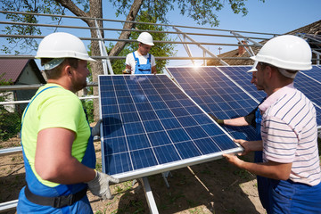 Team of three young technicians in protective helmets adjusting heavy solar photo voltaic panels on...