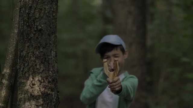 boy hides behind a tree and aims at someone with a slingshot outdoors