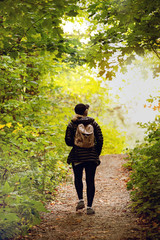 blond woman in dark hiking clothes and pink backpack walking in forest in autumn in park, Seen from behind, telephoto lens. Light effect on sunset. Gatineau Park, Quebec, Ontario.