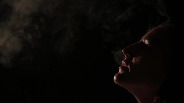 Pensive brunette in a white shirt smokes a hookah in the dark. Portrait of a woman smoking a vape, hovering. Black background. Electronic Cigarette. Bad habit.