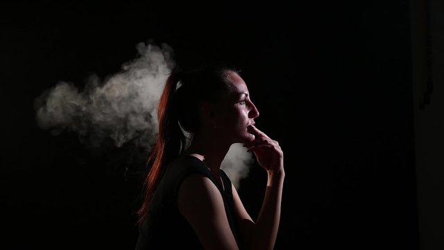 A sad brunette is in a cloud of smoke. Portrait of a woman smoking a vape behind her, soaring. Black background. Passive smoking. Bad habits.