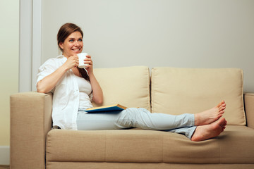 Fototapeta premium Smiling woman drinking coffee sitting on sofa with book.
