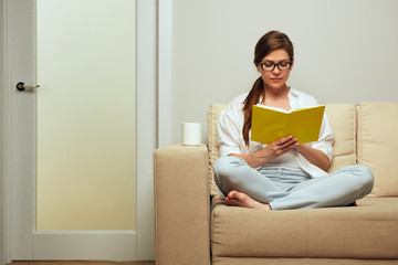 Young woman with glasses sitting on sofa reading book.