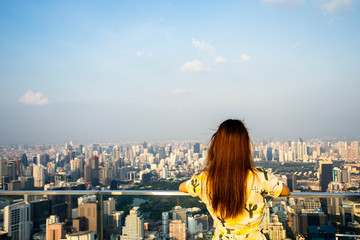 Fototapeta na wymiar Woman wear yellow dress, Asian traveler standing with their backs turned and looking aside on balcony with beautiful modern big city view on background.