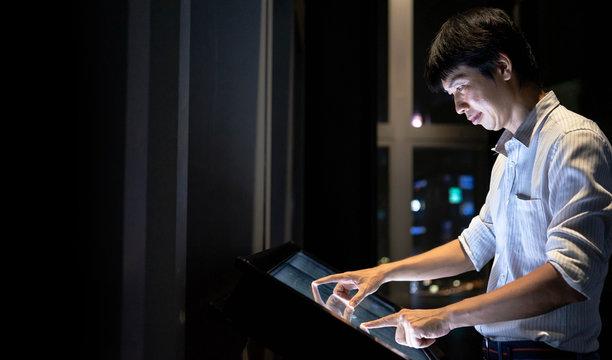 Man Interacting With Large Touch Screen Large Display While Consults About Touristic Information With Copy Space On Black Background. Interactive Kiosk With Public Transport Large Building Map.