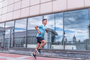 Fototapeta na wymiar male athlete runs in morning, summer, spring in city, training in jump. Active lifestyle, workout, fitness in fresh air. Motivation for sports. Glass windows background. Free space.