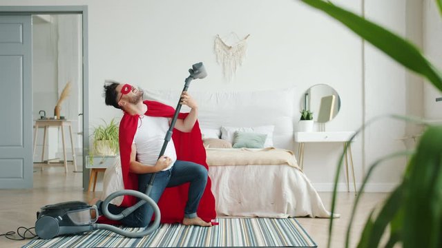 Crazy guy in superman costume is dancing with vacuum cleaner having fun indoors vacuuming carpet in bedroom. Funny people, super hero and lifestyle concept.