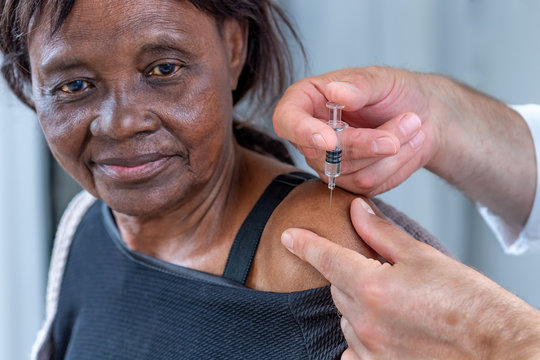 African Senior Woman Receiving Disease Prevention Vaccine, .Caucasian Hands Holding Syringe With Needle Next To Upper Arm