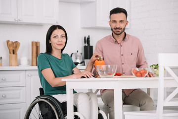 attractive disabled woman with boyfriend smiling at camera while sitting at kitchen table and preparing salad together