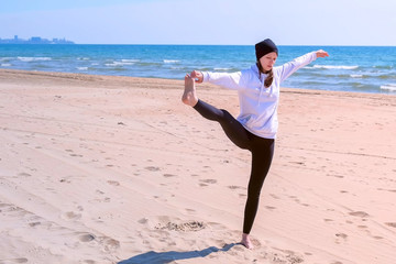 Woman practices yoga on sea beach on sand. She makes exercise raises her leg and holds it hand in...