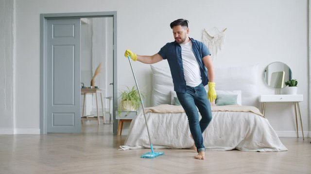 Slow motion of attractive young man in rubber gloves washing wooden floor and dancing with plastic mop having fun in flat. People and housework concept.