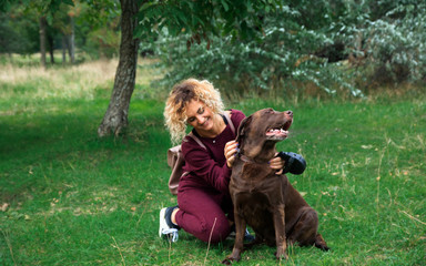 Naklejka na ściany i meble blonde curly girl plays with chocolate labrador in green park