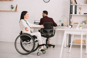 cheerful disabled businesswoman gesturing while sitting in wheelchair near colleague