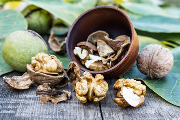 Walnut kernels lie in a bowl next to nuts in green shells and green leaves on a rustic old wooden table.