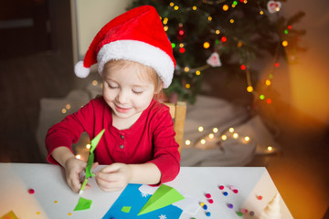 a little girl makes a card application of colored paper on the background of the Christmas tree. the concept of new year kids creativity