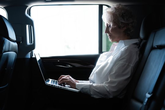 Senior Businesswoman Travelling To Office In A Car Sitting On Backseat With Laptop.