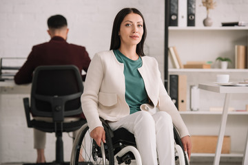 smiling disabled businesswoman looking at camera while sitting in wheelchair near colleague working...